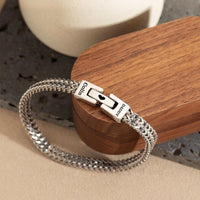 La Pulsera grabada - A mi amor es una cadena plateada con broche rectangular personalizada con nombres y un corazón negro, perfecta como regalo para hombre, presentada sobre madera y piedra.