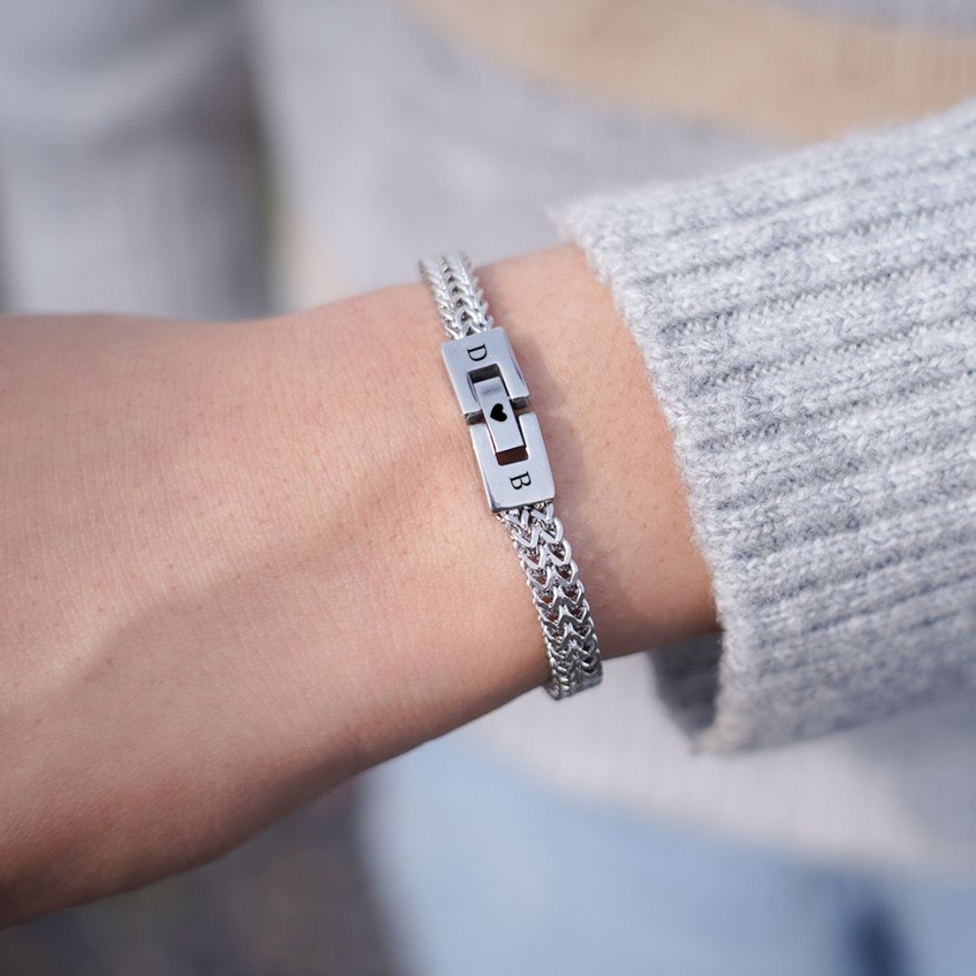 Un primer plano muestra una muñeca que lleva la "Pulsera grabada - A mi amor", una pulsera de cadena de plata con cierre grabado. Combinada con un jersey de punto gris, es el regalo perfecto para hombre.