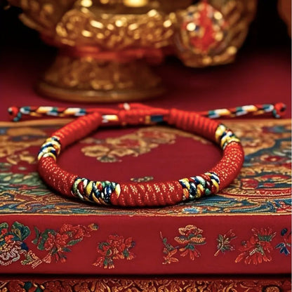 La Pulsera Hilo rojo del destino (Oferta 2X1), trenzada con hilos rojos, amarillos, azules y blancos, descansa sobre una ornamentada caja roja bordada con detalles dorados, ideal para quienes buscan protección energética.