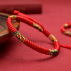 Un primer plano de la Pulsera Hilo rojo del destino (Oferta 2X1), conocida por ofrecer protección energética, con coloridas secciones tejidas. Descansa sobre una superficie roja junto a un cuenco de madera, con una iluminación que resalta su detallada artesanía.