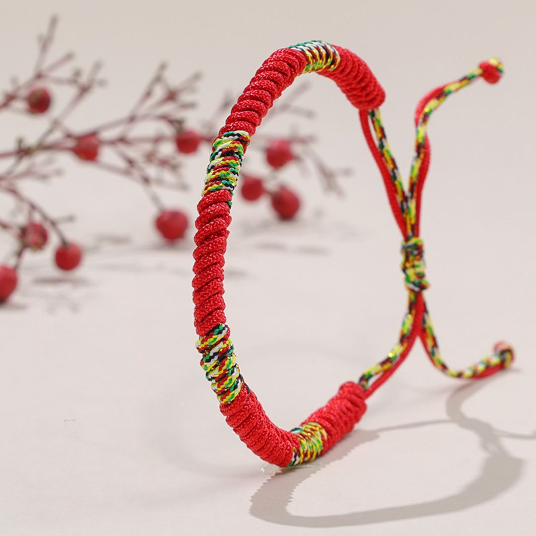 La Pulsera Hilo rojo del destino (Oferta 2X1) se presenta de pie con un nudo suelto; ramas con frutos rojos decoran el fondo claro, destacando esta pulsera como símbolo de suerte y protección energética.