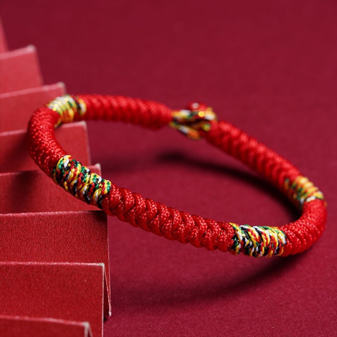 La Pulsera Hilo rojo del destino (Oferta 2X1) luce un trenzado con hilos multicolores, mostrada sobre fondo rojo. Perfecta como amuleto de la suerte y protección energética para usar a diario.
