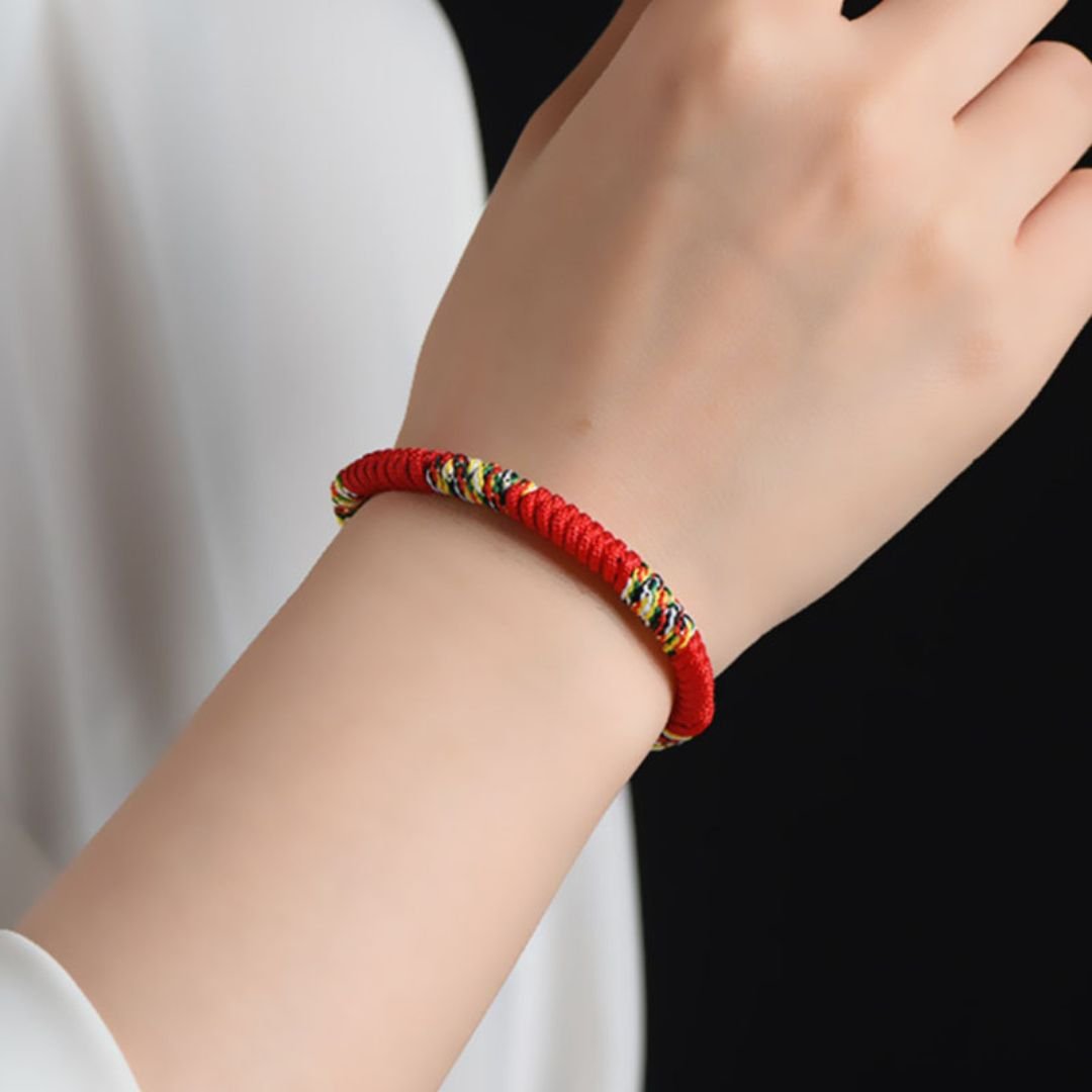 Sobre un fondo negro se muestra una mano que lleva la Pulsera Hilo rojo del destino (Oferta 2X1), trenzada intrincadamente con hilos multicolores. La manga blanca resalta esta pulsera de la suerte y su protección energética.