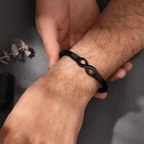 Una persona ajusta cuidadosamente una Pulsera Infinito - A mi hijo en su muñeca. Cerca, unas hojas secas decorativas y una piedra oscura se posan sobre la superficie gris, convirtiéndola en un regalo ideal para hijo.