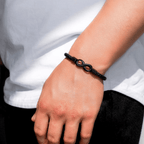 Una persona con la Pulsera Infinito - A mi hijo, una pulsera de cuero negro con un charm infinito negro, combinada con una camisa blanca y unos pantalones negros: un regalo ideal para hijo.