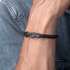 Primer plano de una muñeca que lleva la Pulsera Infinito - A mi hijo, una pulsera de cuero negro con un dije de infinito; regalo perfecto para un hijo. La persona va vestida con una camisa vaquera.
