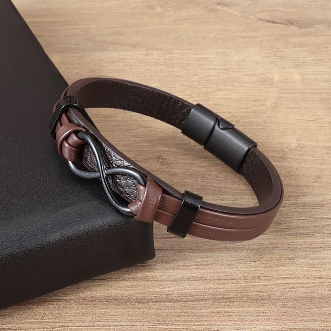 Pulsera Infinito - A mi Hombre: pulsera de cuero para hombre con dije y broche en forma de infinito negro, exhibida sobre madera junto a un objeto rectangular negro, el regalo perfecto para él.