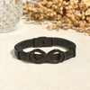 La Pulsera Infinito - A mi Hombre, una elegante pulsera de cuero negro con un charm del símbolo del infinito, se muestra en una superficie clara junto a flores secas y cristal texturizado: un regalo ideal para hombre.