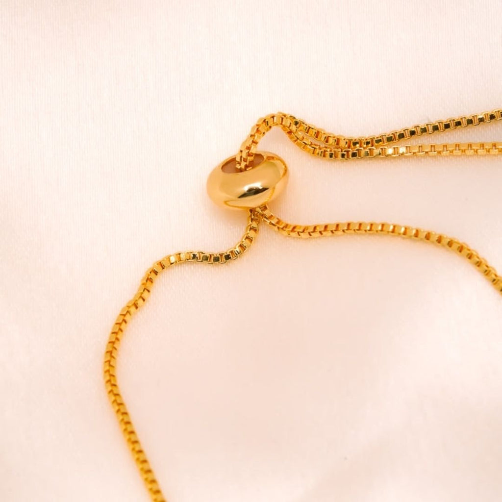 Un primer plano de un collar de cadena dorada con un colgante redondeado sobre tela rosa claro, ideal para combinar con la Pulsera Infinito con ojo turco (OFERTA 2X1) para una protección elegante contra la energía negativa.