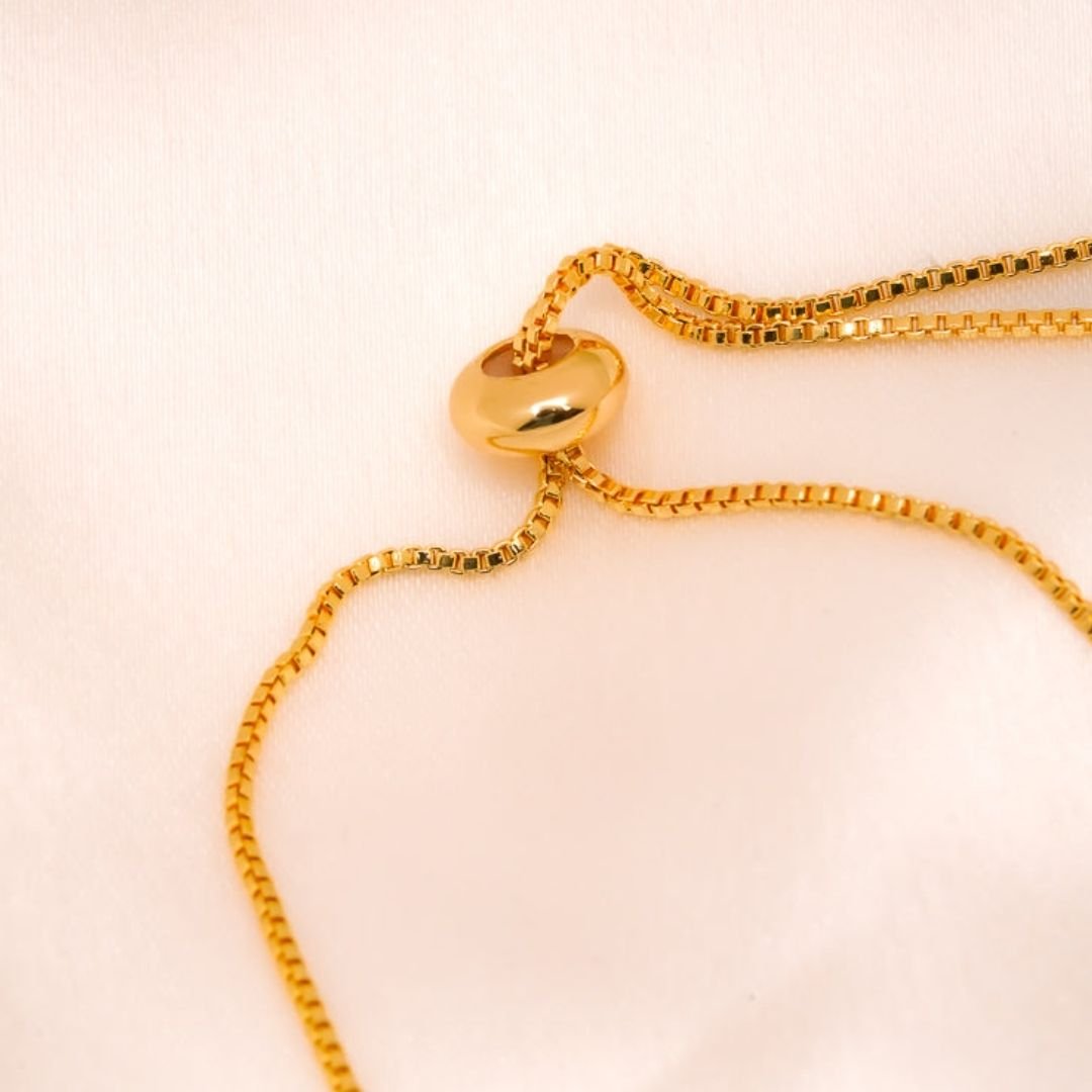 Un primer plano de un collar de cadena dorada con un colgante redondeado sobre tela rosa claro, ideal para combinar con la Pulsera Infinito con ojo turco (OFERTA 2X1) para una protección elegante contra la energía negativa.