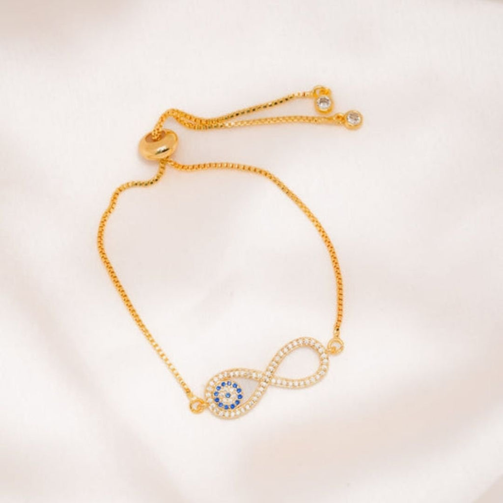 La Pulsera Infinito con ojo turco (OFERTA 2X1) presenta una pulsera de cadena dorada con un símbolo de infinito y piedras azules y blancas, diseñada para proteger contra la energía negativa y el mal de ojo. Se presenta en un tejido suave de color claro.