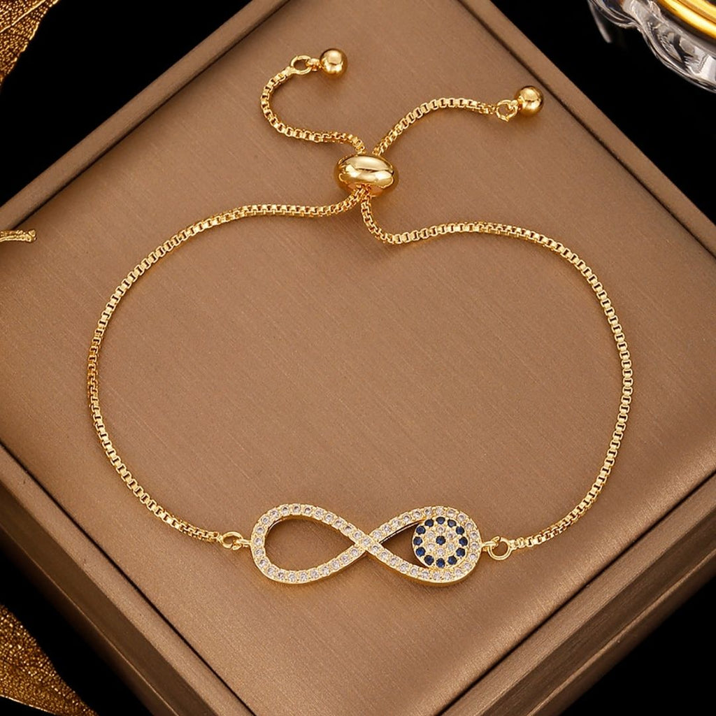 La Pulsera Infinito con ojo turco (OFERTA 2X1) es una pulsera de cadena de oro con un símbolo de infinito, piedras blancas y un racimo de piedras azules para la protección contra la energía negativa, que se muestra sobre un fondo de caja de color tostado.