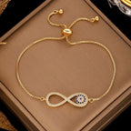 La Pulsera Infinito con ojo turco (OFERTA 2X1) es una pulsera de cadena de oro con un símbolo de infinito, piedras blancas y un racimo de piedras azules para la protección contra la energía negativa, que se muestra sobre un fondo de caja de color tostado.