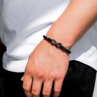 Una persona con camisa blanca y pantalón negro luce su muñeca adornada con la Pulsera Infinito personalizada - A mi Hombre, convirtiéndola en un regalo ideal para hombre.