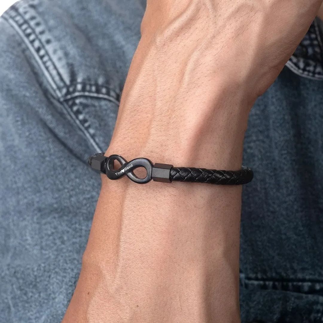 Primer plano de la muñeca de una persona que lleva la Pulsera Infinito personalizada - A mi Hombre, una pulsera de cuero trenzado negro con cierre infinito de metal, contrastada sobre un fondo de chaqueta vaquera azul.