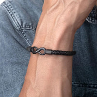 Primer plano de la muñeca de una persona que lleva la Pulsera Infinito personalizada - A mi Hombre, una pulsera de cuero trenzado negro con cierre infinito de metal, contrastada sobre un fondo de chaqueta vaquera azul.
