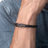 Primer plano de la muñeca de una persona que lleva la Pulsera Infinito personalizada - A mi Hombre, una pulsera de cuero trenzado negro con cierre infinito de metal, contrastada sobre un fondo de chaqueta vaquera azul.