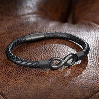 La Pulsera Infinito personalizada - A mi Hombre es una pulsera de cuero trenzado negro con un cierre infinito de metal grabado con texto, desplegado sobre cuero marrón: un regalo ideal para hombre.