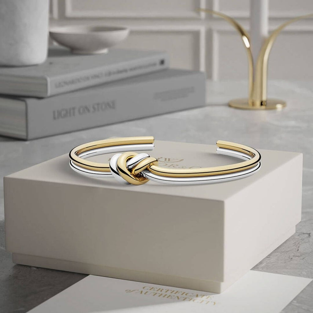 La Pulsera Nudo infinito dual, un brazalete de acero inoxidable con nudos de oro y plata, descansa sobre un joyero blanco con libros y un objeto decorativo sobre una mesa de mármol al fondo.
