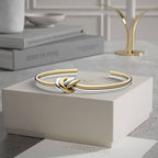La Pulsera Nudo infinito dual, un brazalete de acero inoxidable con nudos de oro y plata, descansa sobre un joyero blanco con libros y un objeto decorativo sobre una mesa de mármol al fondo.