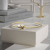 La Pulsera Nudo infinito dual, un brazalete de acero inoxidable con nudos de oro y plata, descansa sobre un joyero blanco con libros y un objeto decorativo sobre una mesa de mármol al fondo.