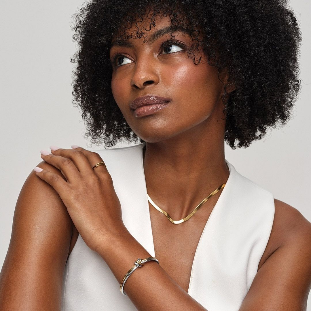 Una mujer de pelo rizado lleva un top blanco sin mangas y joyas de oro, entre ellas la Pulsera Nudo infinito dual, un collar y un anillo. Mirando hacia arriba con la mano sobre el hombro, irradia elegancia: un look ideal para un regalo de aniversario.