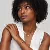 Una mujer de pelo rizado lleva un top blanco sin mangas y joyas de oro, entre ellas la Pulsera Nudo infinito dual, un collar y un anillo. Mirando hacia arriba con la mano sobre el hombro, irradia elegancia: un look ideal para un regalo de aniversario.