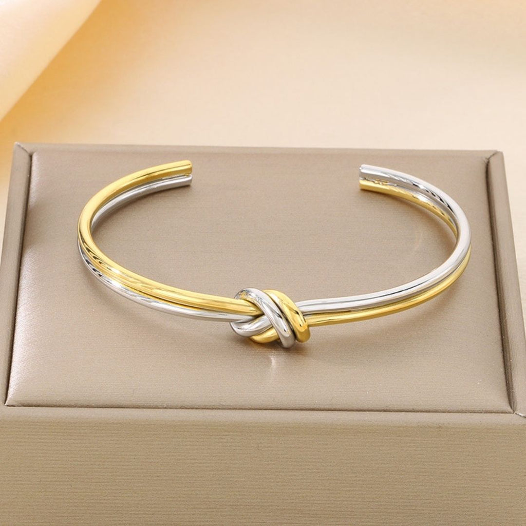 La Pulsera Nudo infinito dual es un brazalete abierto bicolor dorado y plateado de acero inoxidable con un diseño de nudo central, elegantemente presentado en una caja acolchada de color beige.