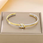 La Pulsera Nudo infinito dual es un brazalete abierto bicolor dorado y plateado de acero inoxidable con un diseño de nudo central, elegantemente presentado en una caja acolchada de color beige.