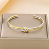 La Pulsera Nudo infinito dual es un brazalete abierto bicolor dorado y plateado de acero inoxidable con un diseño de nudo central, elegantemente presentado en una caja acolchada de color beige.