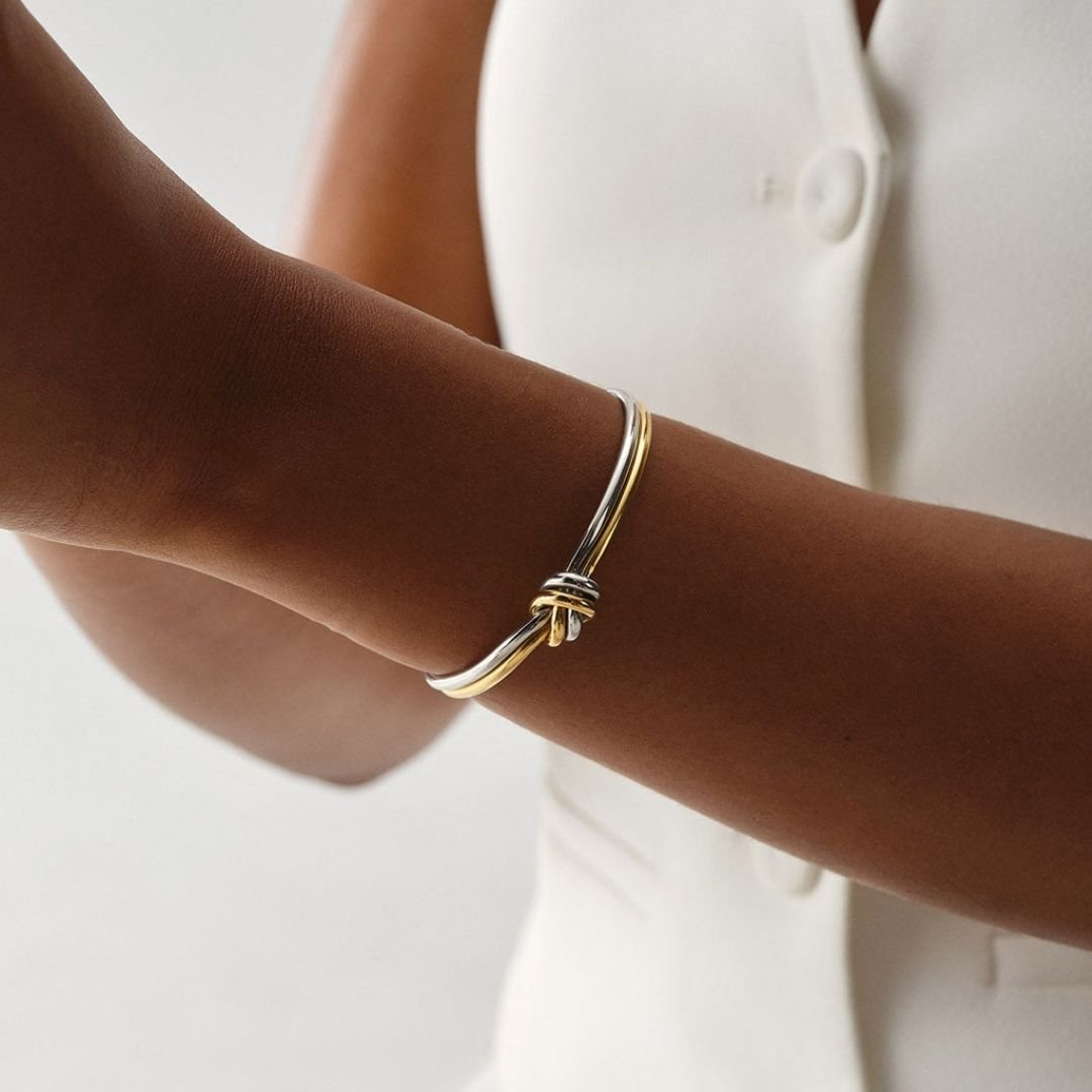 Primer plano del brazo de una persona que lleva la Pulsera Nudo infinito dual, un brazalete unisex de acero inoxidable bicolor dorado y plateado, elegantemente combinado con un top blanco abotonado sin mangas, ideal como regalo aniversario.