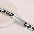 La Pulsera personalizada con frase para hombre, en plata y negro de acero inoxidable, lleva el mensaje grabado "Mensaje grabado" en la placa frontal; se muestra sobre una superficie circular clara y es perfecta como regalo para hombre.
