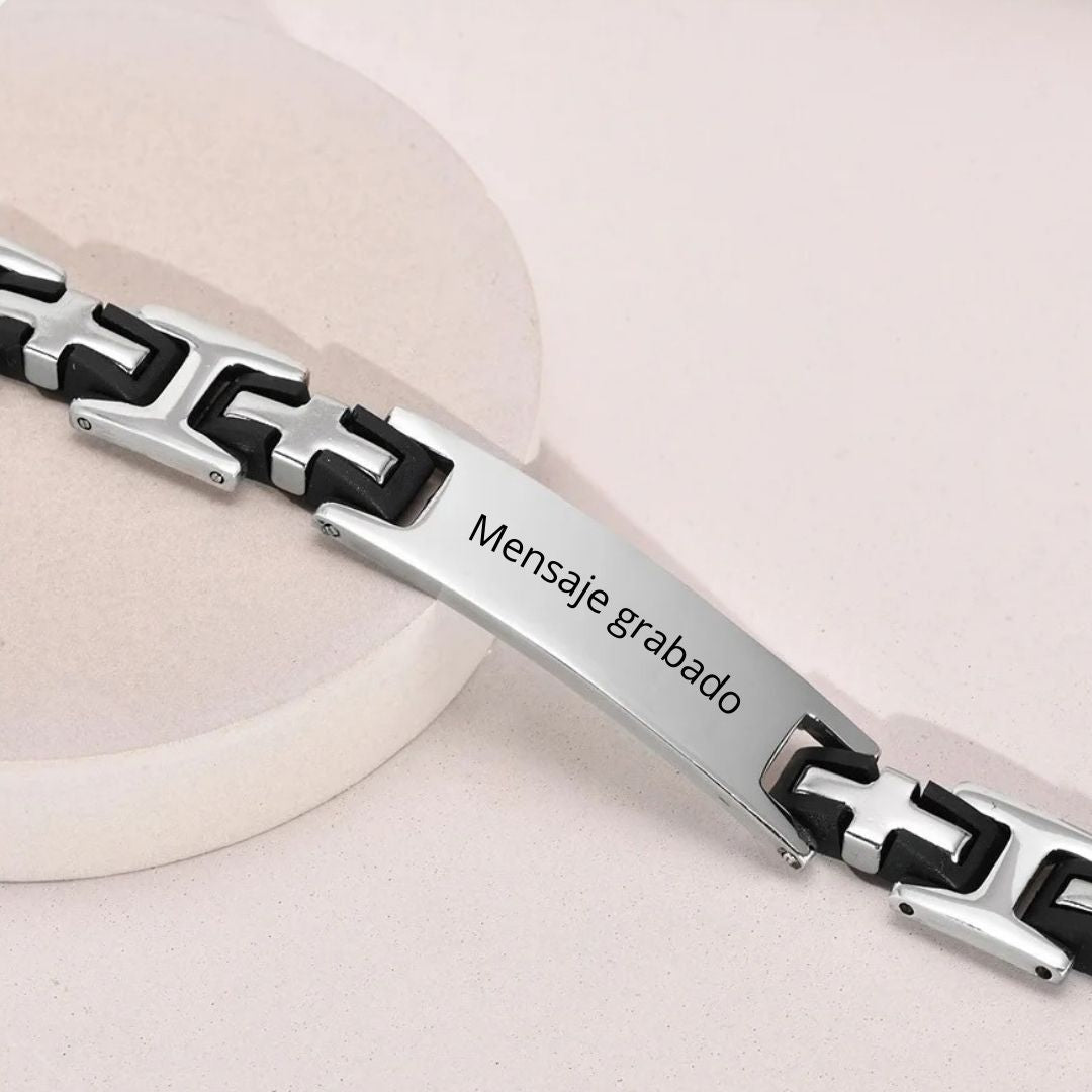 La Pulsera personalizada con frase para hombre, en plata y negro de acero inoxidable, lleva el mensaje grabado "Mensaje grabado" en la placa frontal; se muestra sobre una superficie circular clara y es perfecta como regalo para hombre.