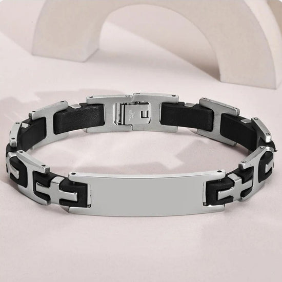 La Pulsera personalizada con frase para hombre combina eslabones en negro y plateado de acero inoxidable, con una placa rectangular central. Se muestra sobre superficie clara y fondo blanco curvo.