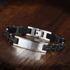 Pulsera personalizada con frase para hombre: brazalete de acero inoxidable y eslabones negros, placa rectangular plateada y diseño industrial. Ideal como regalo para hombre, presentada sobre superficie de madera pulida.