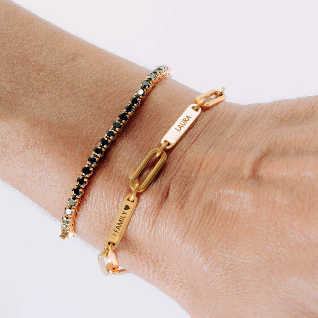 Una muñeca con dos pulseras: una Pulsera personalizada con nombres, cadena de oro grabada con FAMILY ♥ y LAURA, y una fina pulsera de cuentas negras y doradas-perfecta como regalo para mamá.