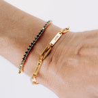 Una muñeca con dos pulseras: una Pulsera personalizada con nombres, cadena de oro grabada con FAMILY ♥ y LAURA, y una fina pulsera de cuentas negras y doradas-perfecta como regalo para mamá.