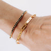 Una muñeca con dos pulseras: una Pulsera personalizada con nombres, cadena de oro grabada con FAMILY ♥ y LAURA, y una fina pulsera de cuentas negras y doradas-perfecta como regalo para mamá.