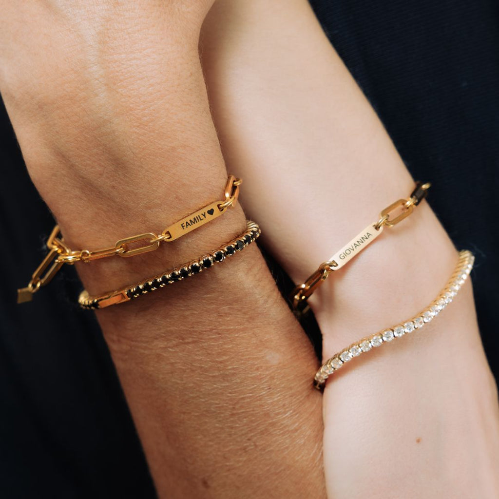 Dos muñecas llevan pulsera de oro personalizada con nombres: una de eslabones con "FAMILY ♥" y nombres grabados, otra con "GIOVANNI", además de una pulsera de cuentas y una pulsera tenis de oro brillante con piedras transparentes.