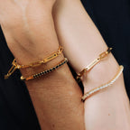 Dos muñecas llevan pulsera de oro personalizada con nombres: una de eslabones con "FAMILY ♥" y nombres grabados, otra con "GIOVANNI", además de una pulsera de cuentas y una pulsera tenis de oro brillante con piedras transparentes.