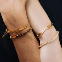 Dos muñecas llevan pulsera de oro personalizada con nombres: una de eslabones con "FAMILY ♥" y nombres grabados, otra con "GIOVANNI", además de una pulsera de cuentas y una pulsera tenis de oro brillante con piedras transparentes.