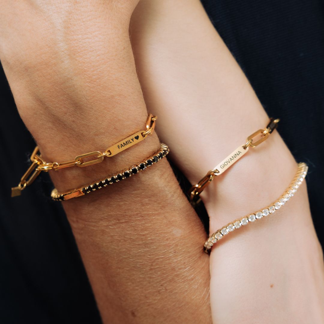 Dos muñecas llevan pulsera de oro personalizada con nombres: una de eslabones con "FAMILY ♥" y nombres grabados, otra con "GIOVANNI", además de una pulsera de cuentas y una pulsera tenis de oro brillante con piedras transparentes.