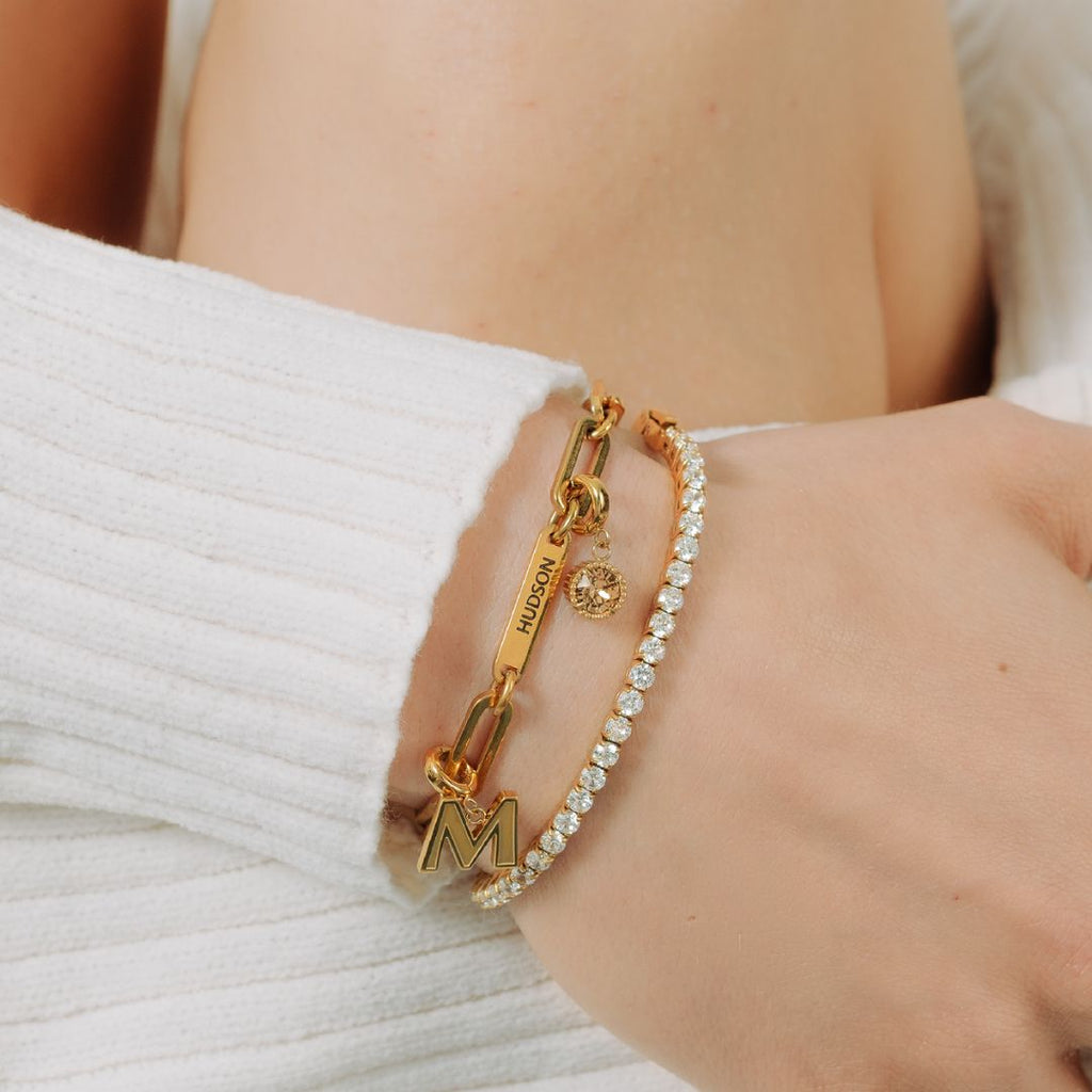 Primer plano de una muñeca con dos pulseras de oro: una Pulsera personalizada con nombres con un dije "M" y "HUDSON", y una delicada pulsera de tenis. La persona lleva un jersey blanco de punto de canalé.