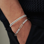 Primer plano de una mano en un bolsillo, que lleva dos pulseras de plata: una enrollada y la otra una pulsera personalizada con nombres. La persona está vestida con pantalones oscuros y un jersey azul marino con textura.