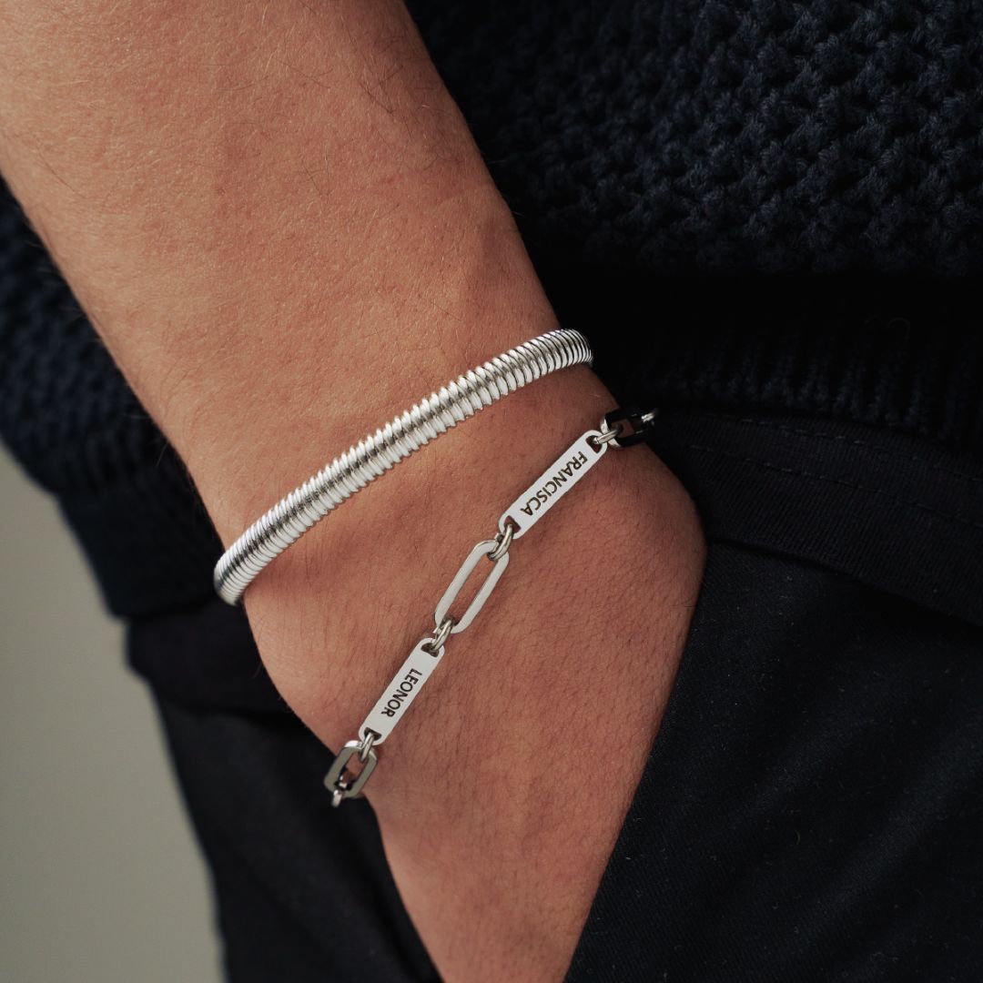 Primer plano de una mano en un bolsillo, que lleva dos pulseras de plata: una enrollada y la otra una pulsera personalizada con nombres. La persona está vestida con pantalones oscuros y un jersey azul marino con textura.