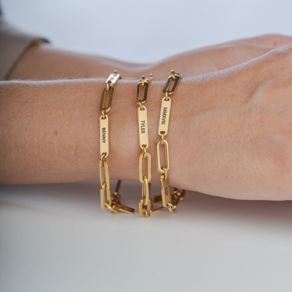 Un primer plano muestra una mano que lleva tres Pulseras personalizadas con los nombres "BRANT", "TYLER" y "MAKENNA" grabados en cadenas de oro. La mano descansa sobre una superficie clara, lo que la convierte en un regalo ideal para mamá.