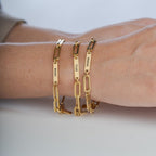 Un primer plano muestra una mano que lleva tres Pulseras personalizadas con los nombres "BRANT", "TYLER" y "MAKENNA" grabados en cadenas de oro. La mano descansa sobre una superficie clara, lo que la convierte en un regalo ideal para mamá.