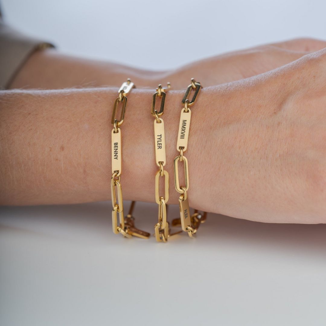 Un primer plano muestra una mano que lleva tres Pulseras personalizadas con los nombres "BRANT", "TYLER" y "MAKENNA" grabados en cadenas de oro. La mano descansa sobre una superficie clara, lo que la convierte en un regalo ideal para mamá.