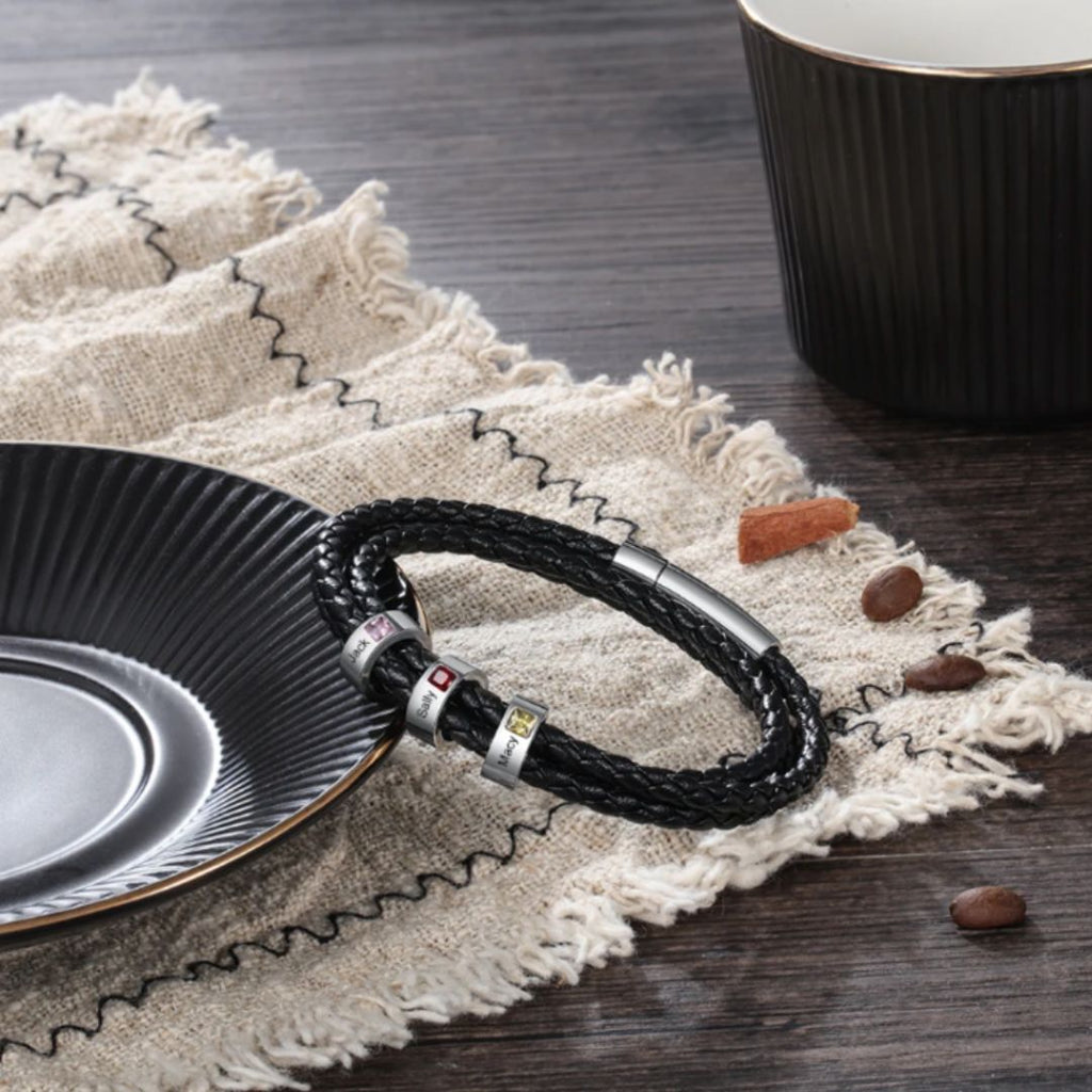 La Pulsera Piedras de vida - A mi Padre, una pulsera de cuero trenzado negro con detalles plateados, descansa sobre una tela beige con flecos cerca de granos de café y canela, lo que la convierte en un regalo ideal Día del Padre.