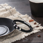 La Pulsera Piedras de vida - A mi Padre, una pulsera de cuero trenzado negro con detalles plateados, descansa sobre una tela beige con flecos cerca de granos de café y canela, lo que la convierte en un regalo ideal Día del Padre.